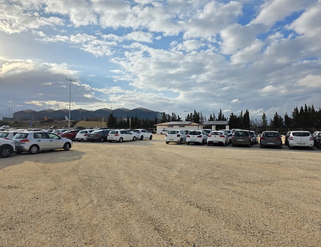 Duży parking z wieloma zaparkowanymi samochodami na utwardzonym placu, w tle góry, drzewa i częściowo zachmurzone niebo.