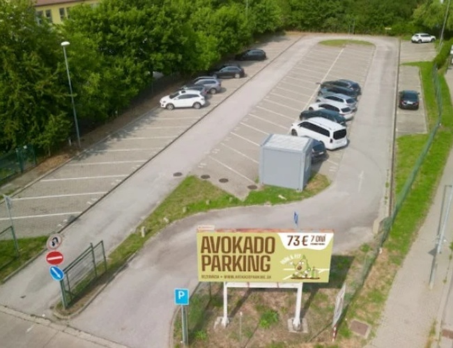 Tablica reklamowa „Avokado Parking” przy wjeździe na parking, obok parkingu i zaparkowanych samochodów, w słoneczny dzień.