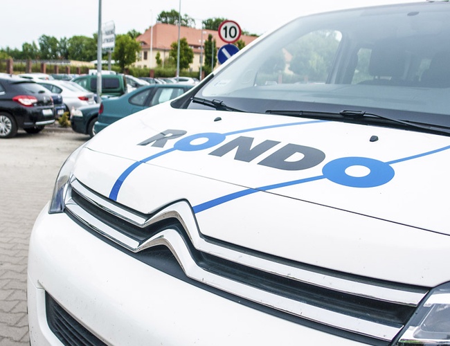 Parking Rondo - na pierwszym planie widać samochód z logo. W tle parkują inne pojazdy. Znaki wskazują ograniczenie prędkości.