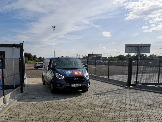 Parking A1 Rondo - Ogrodzony plac z utwardzoną nawierzchnią. Na pierwszym planie stoi oznakowany bus. W tle widoczny billboard.