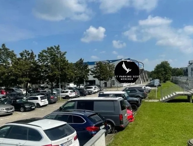 Zewnętrzny parking pełen samochodów w słoneczny dzień, z budynkiem i zielonym trawnikiem po lewej stronie.