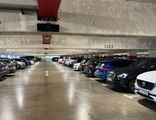 Wnętrze parkingu wielopoziomowego z szeroką drogą przejazdową i samochodami zaparkowanymi po obu stronach. Nad miejscami postojowymi widoczne są elementy konstrukcji stropu i oświetlenie.