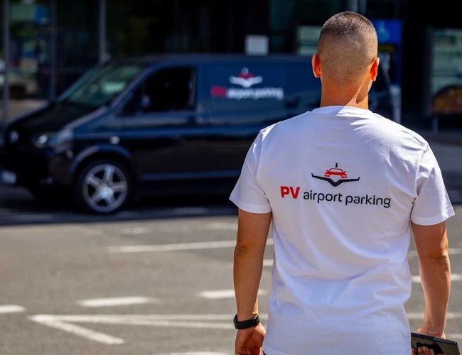 Pracownik PV Airport Parking spacerujący po terenie parkingu lotniskowego, widoczny na tle samochodu transferowego z oznaczeniem parkingu.