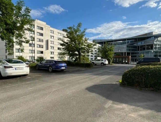 Supreme Parkservice – einige Autos stehen auf einem großen Platz. Der Parkplatz ist von einer Hecke umgeben. Im Hintergrund befinden sich Gebäude.