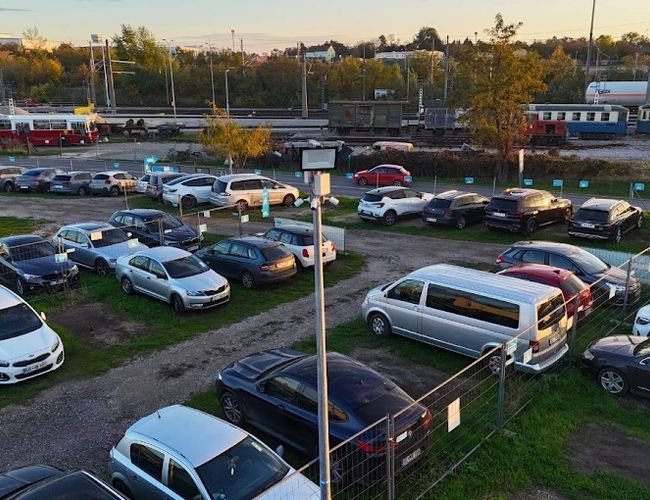 Parktiger Airport Parking 1 - Na parkingu stoją samochody na trawiastej nawierzchni. Teren jest ogrodzony i dobrze oświetlony. W tle widać tory kolejowe.