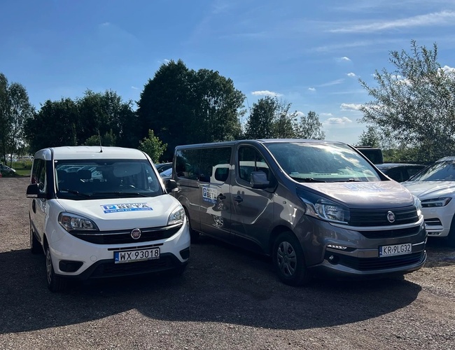 Parking Beta - En el área de grava hay dos furgonetas. El terreno está rodeado de vegetación exuberante y árboles. El clima es soleado con algunas nubes.