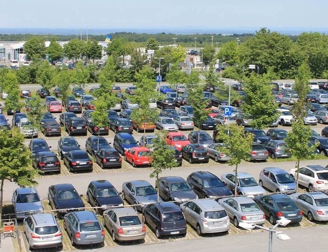 Widok z góry na rozległy parking terenowy z licznymi zaparkowanymi samochodami. Parking otoczony jest drzewami i zielenią.