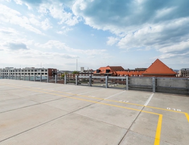 Parkovisko Best Valet Park Service - je viditeľná betónová strecha parkoviska. Na oblohe sú vidieť budovy s červenými strechami. Okraj parkoviska je ohradený.