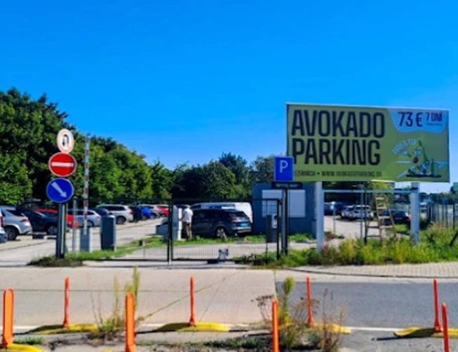 Wjazd na „Avokado Parking” z widoczną tablicą reklamową, znakami drogowymi i barierkami zabezpieczającymi przy jezdni.