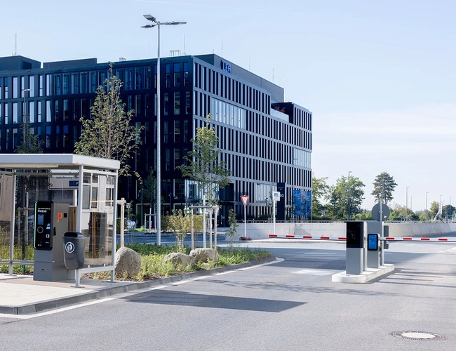 Wejście na parking P25 z infrastrukturą dla pieszych, drogą dojazdową oraz nowoczesnym budynkiem w tle.