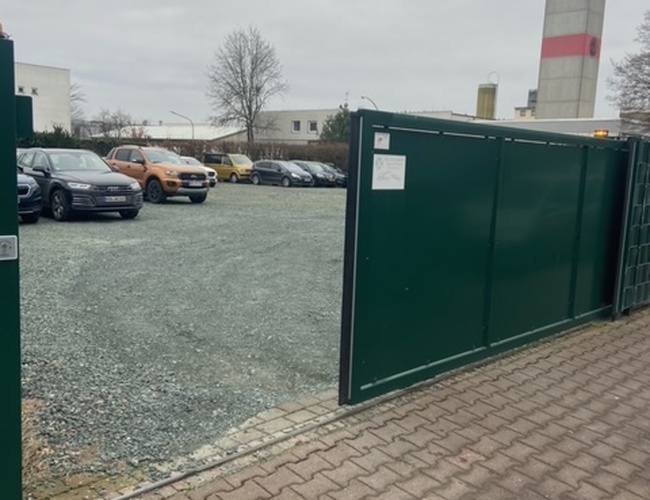 Estacionamiento Park-Box-Frankfurt Valet - El estacionamiento está cercado con una valla de metal. Varios coches están aparcados en el suelo de grava. En el fondo se puede ver un edificio.