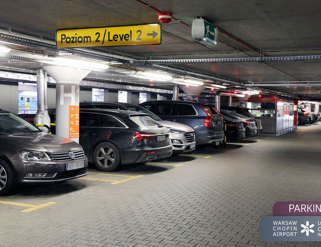 Oficjalny Parking Lotniska P2 – wewnętrzny parking wielopoziomowy. Kilka samochodów zaparkowanych obok siebie. Na suficie znaki kierunkowe.