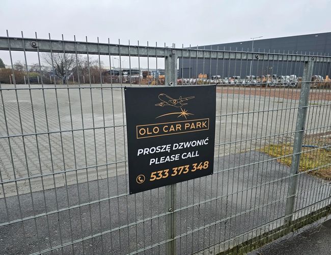 Parking Olo Car Park - Teren jest ogrodzony metalowym płotem. Na bramie widoczna jest tablica informacyjna z numerem telefonu. W tle stoją samochody oraz magazyn.