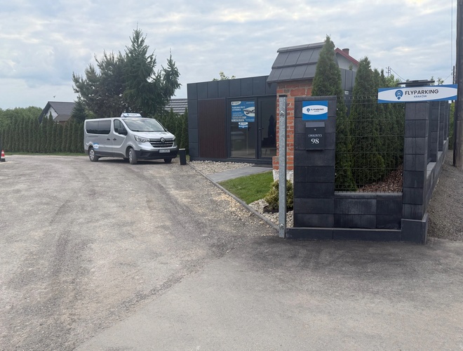 FlyParking – Przed budynkiem stoi zaparkowany biały bus. Ogrodzenie jest murowane. Obok rosną tuje.