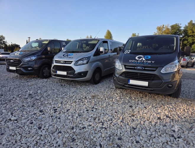Odlotowy Parking Mado - Dwa busy stoją przed zamkniętą bramą. W tle drzewa i zachmurzone niebo.