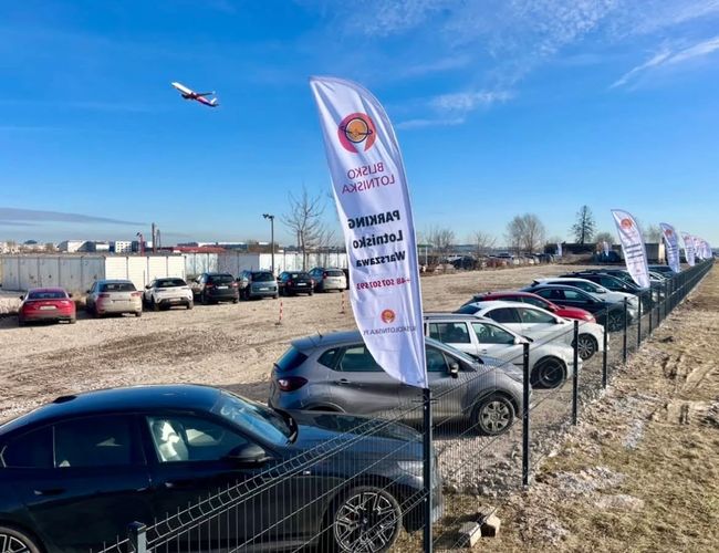 Parking Blisko Lotniska - Ogrodzony teren z zaparkowanymi samochodami. Na niebie leci samolot. Zimowy krajobraz i błękitne niebo.