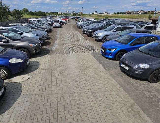 Estacionamiento U Madzi - Filas de coches estacionados a lo largo del camino de piedra. El área está cercada y iluminada.