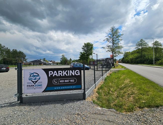ParkZone – Ogrodzony parking z żwirem. Po lewej kilka aut. Obok parkingu biegnie droga. Drzewa otaczają teren.