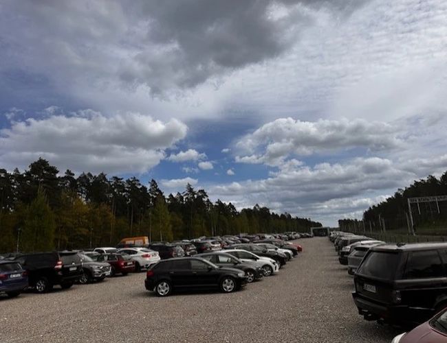 Auto Airport Parking – Na żwirowym placu stoją zaparkowane samochody. Teren otaczają drzewa i jest dobrze oświetlony.