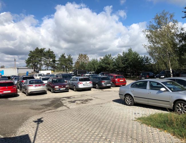Parking A1 – na utwardzonym placu stoją samochody. W tle drzewa osłaniają teren. Na niebie są białe chmury.
