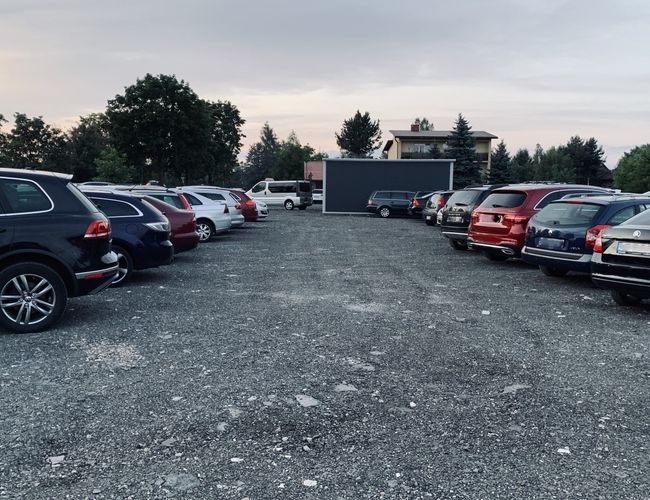 Balice Premium Park - Samochody stoją na nieutwardzonej powierzchni. Parking jest otoczony drzewami. W tle widać budynek.