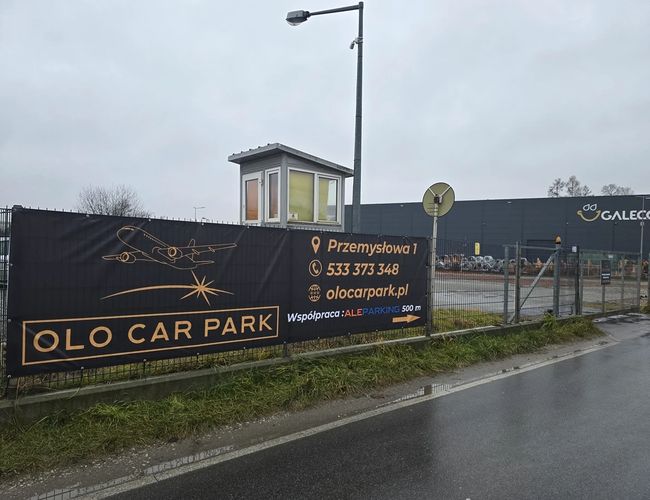 Parking Olo Car Park – ogrodzony teren z banerem reklamowym. W tle jest budka ochrony i szare budynki. Pogoda jest pochmurna.