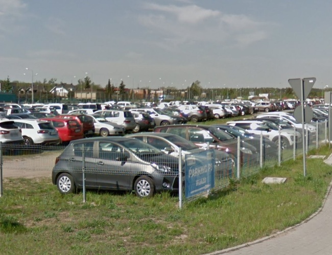 Oficjalny Parking P5/P6 – Ogrodzony teren wypełniony samochodami. Widoczne znaki drogowe i chodnik obok.