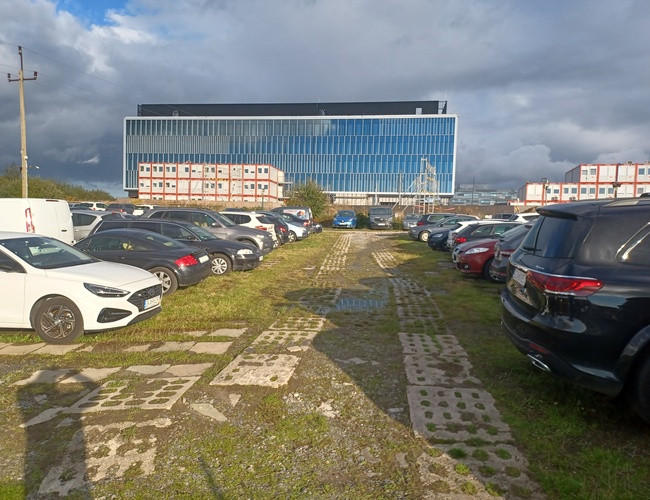 Maxi Parking – Hay muchos coches en la hierba. Al fondo hay un gran edificio de vidrio. A su lado hay contenedores blancos más pequeños.