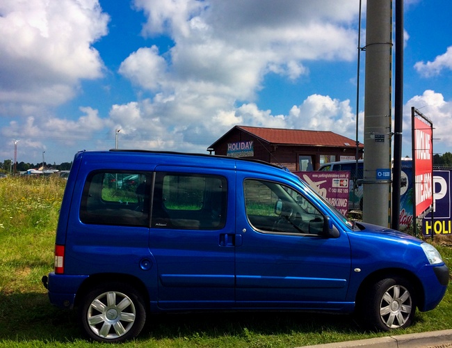 Parkovanie Mega Park Service – Tráva pri okraji vozovky. Stojí modré dodávka. V pozadí budova a oblaky na oblohe.