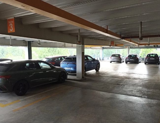 Półotwarty parking z miejscami postojowymi pod zadaszeniem. Samochody zaparkowane są wzdłuż drogi przejazdowej, a na końcu parkingu widoczne jest światło dzienne.