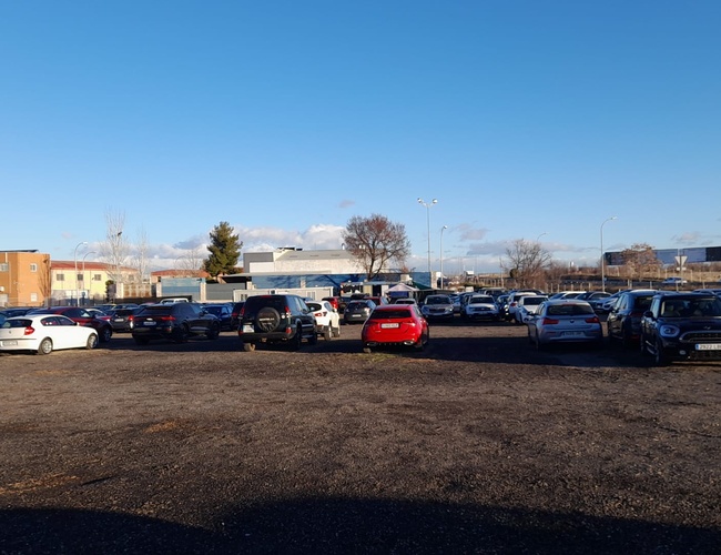 Parkovisko Pleyades Valet - Neuzačlenený povrch plného áut. Na konci stoja niekoľko budov. Na pozadí je modrá obloha.