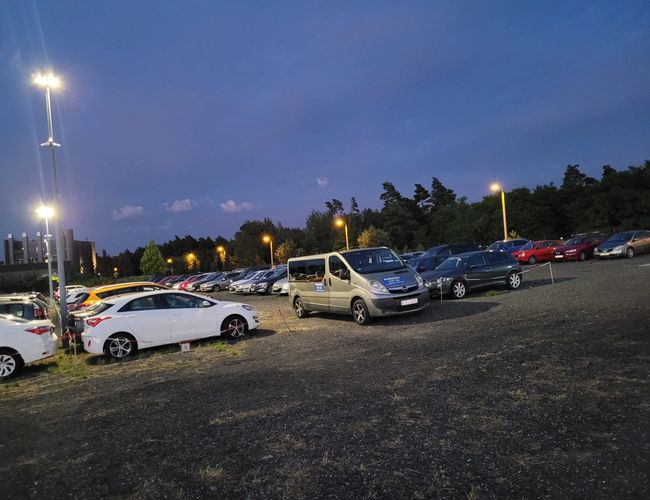 Parking LOT 24 - Na parkingu stoi kilka samochodów. Teren jest dobrze oświetlony latarniami. W tle widać drzewa i budynek.