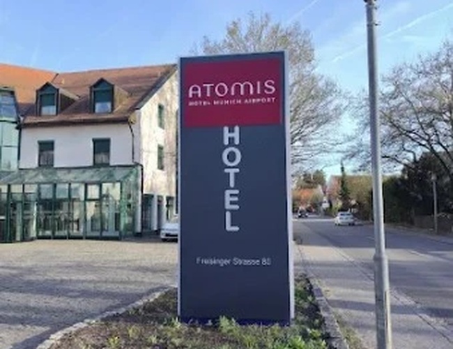 Atomis Hotel Munich Airport Parking Cubierto - Cartel con el nombre del hotel en la calle. Al fondo se ven edificios y árboles.