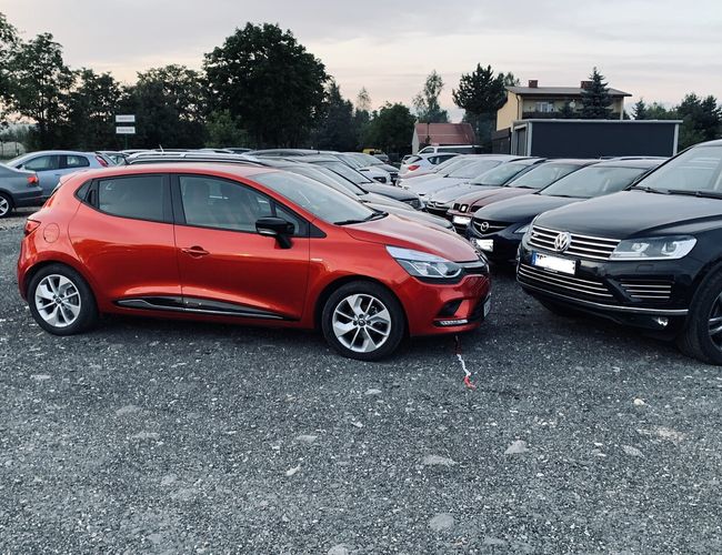 Kraków KING - Na żwirowym parkingu znajduje się wiele samochodów. Teren otaczają drzewa i budynki. Na pierwszym planie jest czerwone auto.