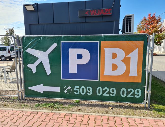 B1 Parking – Stoi tablica z dużym napisem i strzałką. Obok widać kontener i zaparkowane auto. Drzewa w tle mają kolorowe liście.