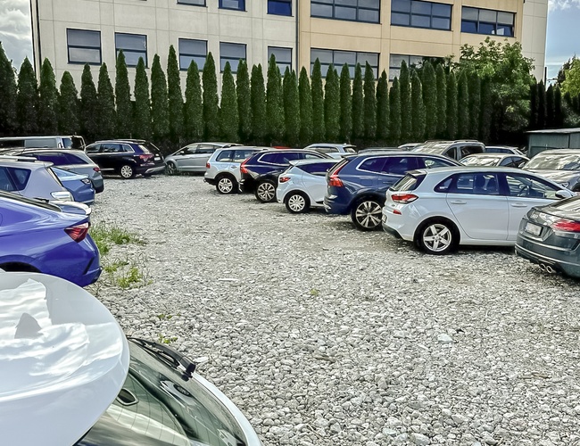 Parksist - štrkový parkovisko s autami pri kancelárskej budove. Pozemok je obklopený ihličnatými stromami. Budova má veľké okná.