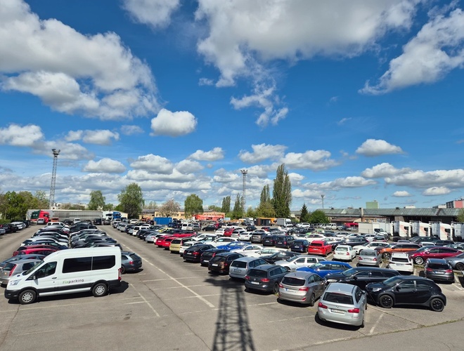 FixParking - Große Parkfläche mit vielen Autos. Das Gelände ist offen und beleuchtet. Im Hintergrund sind Bäume und Wolken sichtbar.