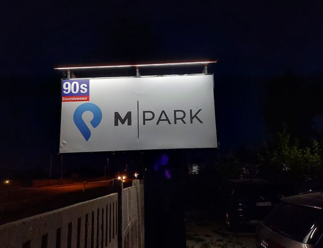 Parking MPark - Señal iluminada visible con el logo de MPark. Entorno nocturno, de fondo un aparcamiento y coches aparcados.