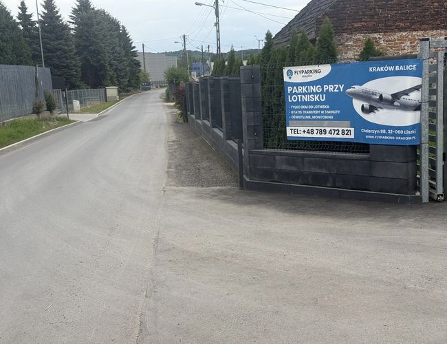 FlyParking – Teren parkingu ogrodzony wysokim płotem. Na bramie wisi baner informacyjny z samolotem.
