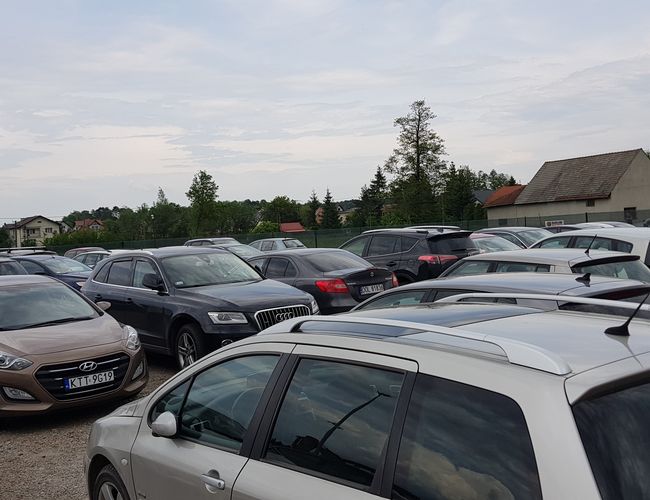 BaliceLot24 - Na parkingu stoi wiele zaparkowanych samochodów. W tle widoczne są drzewa i budynki. Teren jest otwarty.