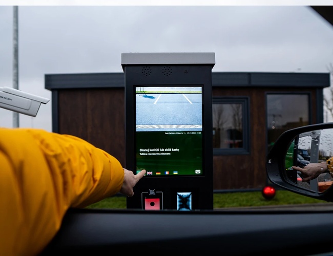 Štandardné jednoduché parkovanie – Osoba dotýka sa obrazovky parkovacieho terminálu. Na pozadí je vidieť budovu s oknami. Vedľa je sledovacia kamera.