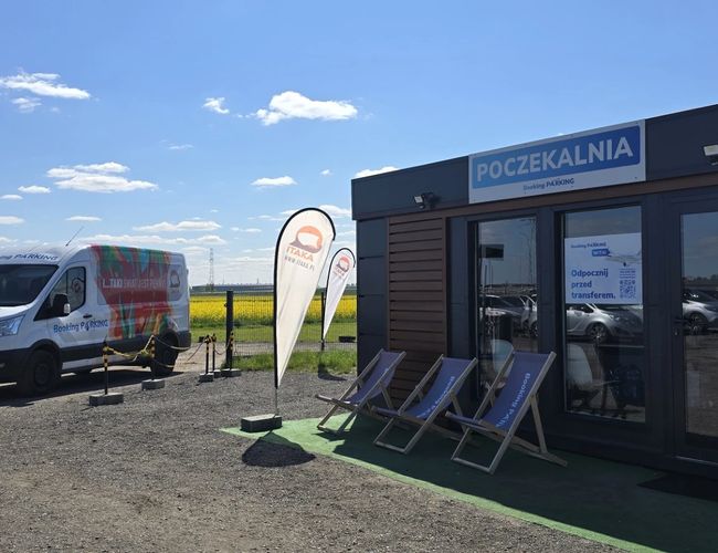 Parking Okęcie Południe – Na zdjęciu widać poczekalnię. Przed nią stoją leżaki i furgonetka. Obok widać flagi reklamowe.
