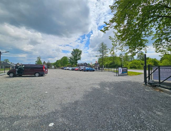 ParkZone – Duży, żwirowy parking z ogrodzeniem. Na miejscu stoją samochody. W oddali widać drzewa i niebo.