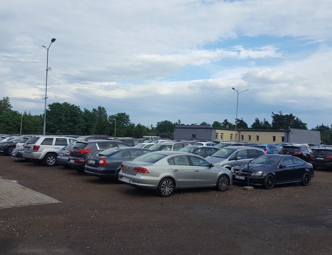 AAP-66 – Parking zapełniony samochodami. Teren nie jest ogrodzony. Widać latarnie i niskie budynki w tle.