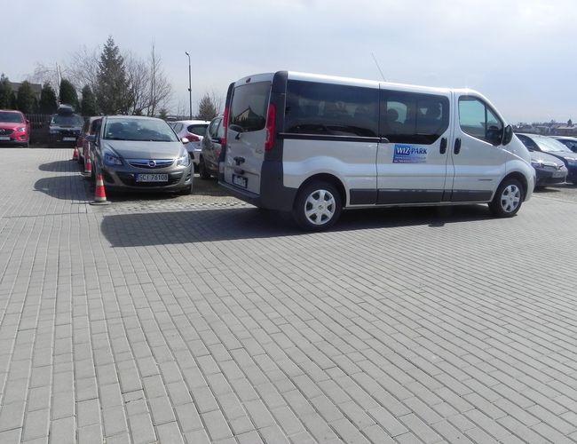 WIZ-PARK – Hay coches en el aparcamiento. El terreno está pavimentado con adoquines. Al fondo se ven árboles y una valla.