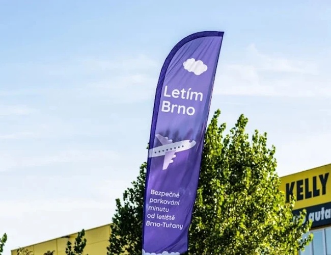 Parking Letim Brno II – na zdjęciu widoczna jest fioletowa flaga reklamowa. Znajdują się na niej napisy informacyjne oraz grafika samolotu. W tle widać drzewo i fragment budynku.
