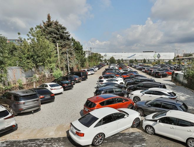 Smart Supreme Parking - Parking pełen samochodów z różnych stron. Teren jest żwirowy i otoczony drzewami. Pogoda jest pochmurna.