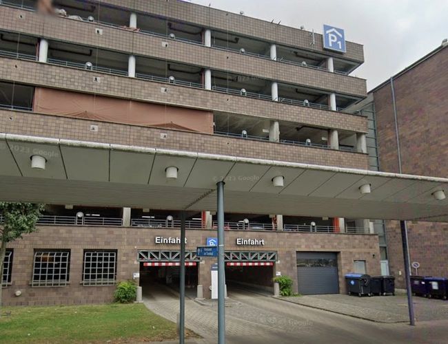 Widok na wielopoziomowy parking od strony wejścia. Przed budynkiem znajduje się zadaszenie nad strefą wejściową oraz oznakowanie parkingu.