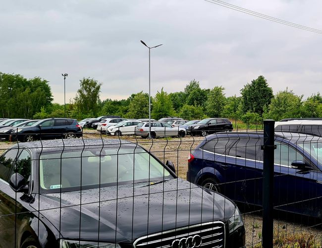 Krispit Parking 24 – samochody stoją za ogrodzeniem. Teren otoczony jest siatką i oświetlony latarniami. W tle widać drzewa.