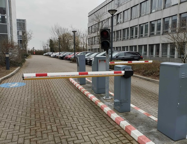 Sun Park Airport Parking Outdoor – Na parkingu są szlabany i sygnalizacja świetlna. W tle stoją zaparkowane samochody. Teren jest otoczony budynkami.
